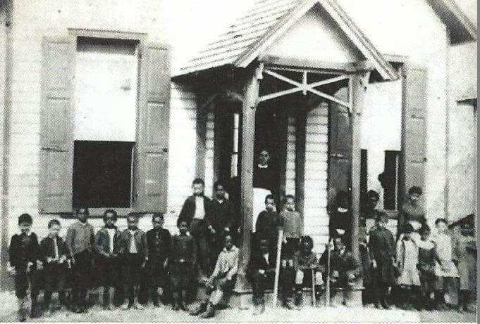A historic black-and-white photo of children and adults standing outside a schoolhouse building