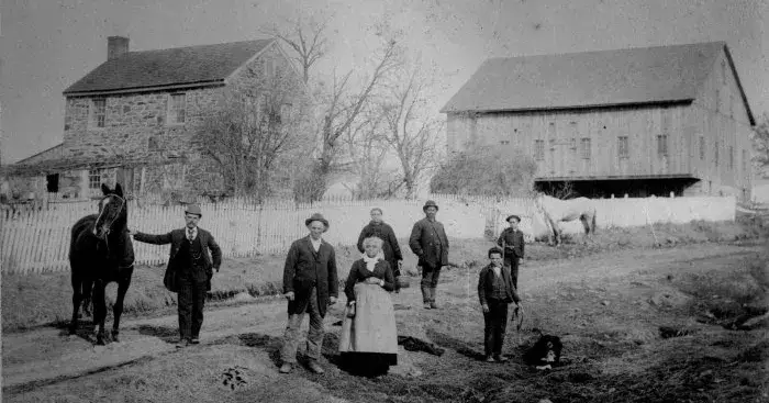 A group of people poses on a dirt path in front of a stone house, a barn, and a picket fence