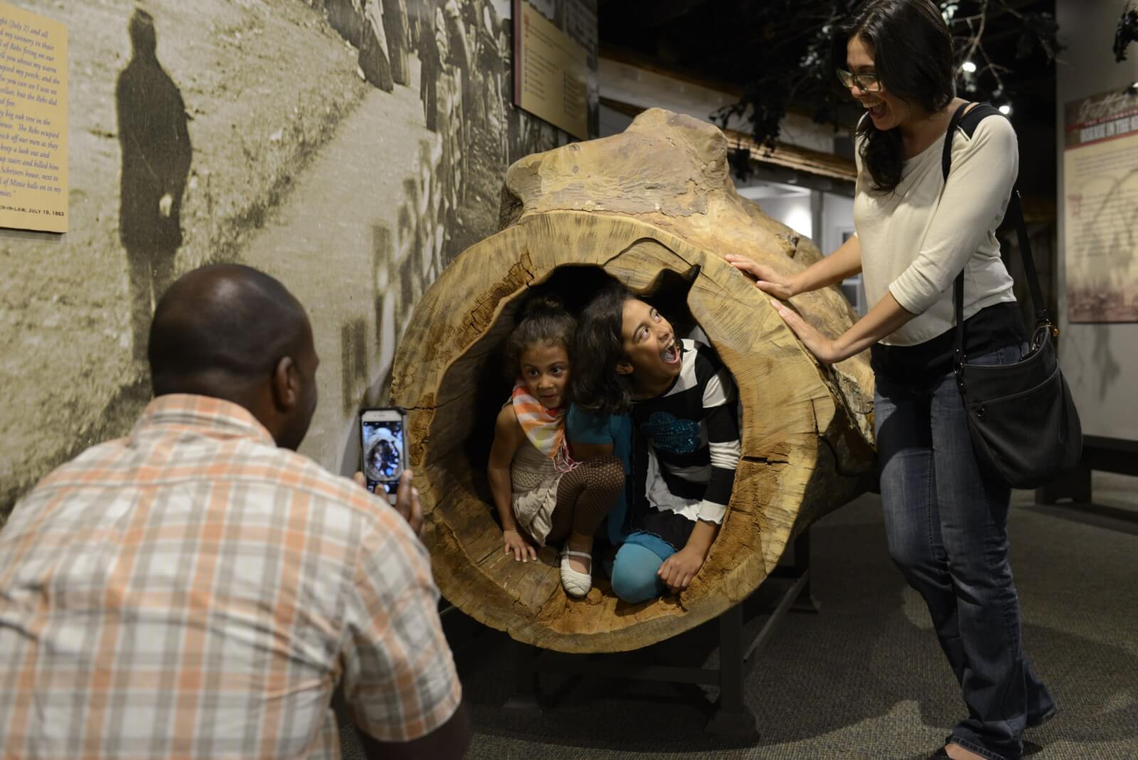 A man takes a phone photo of two young girls posing inside a hollow log display at a museum