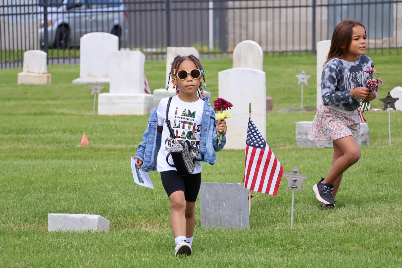 Two young girls carry flowers through a cemetery with white headstones and American flags
