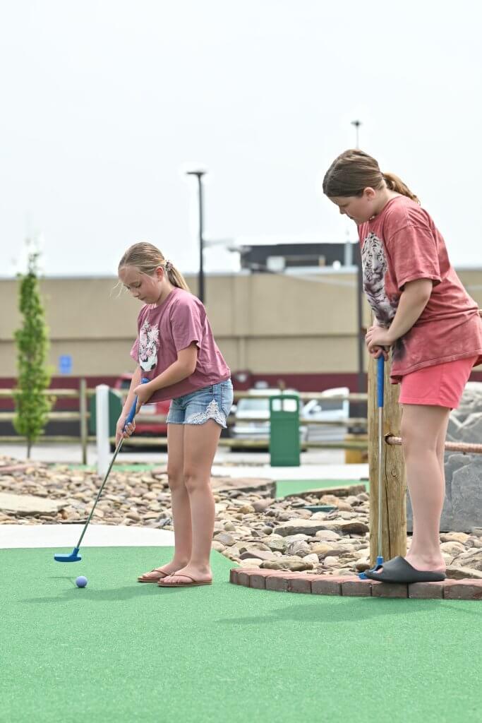 Two young girls play miniature golf together on a green course on a bright day