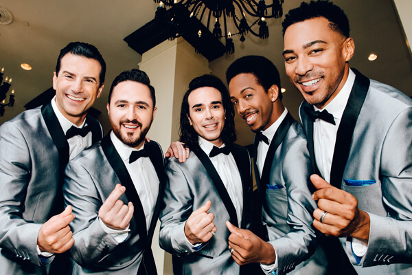 Five guys dressed in silver tuxedo jackets leaning down to the camera while snapping their fingers