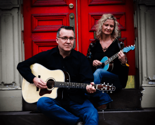 A man and a woman sit on steps in front of a red door playing acoustic stringed instruments