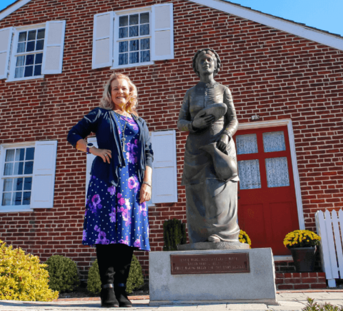 A woman in a floral dress poses next to a bronze statue of a woman in front of a brick house